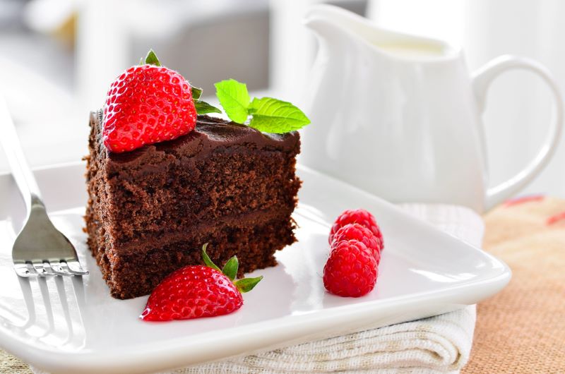 A white plate showing a slice of low FODMAP Chocolate Fudge Cake decorated with strawberries and raspberries