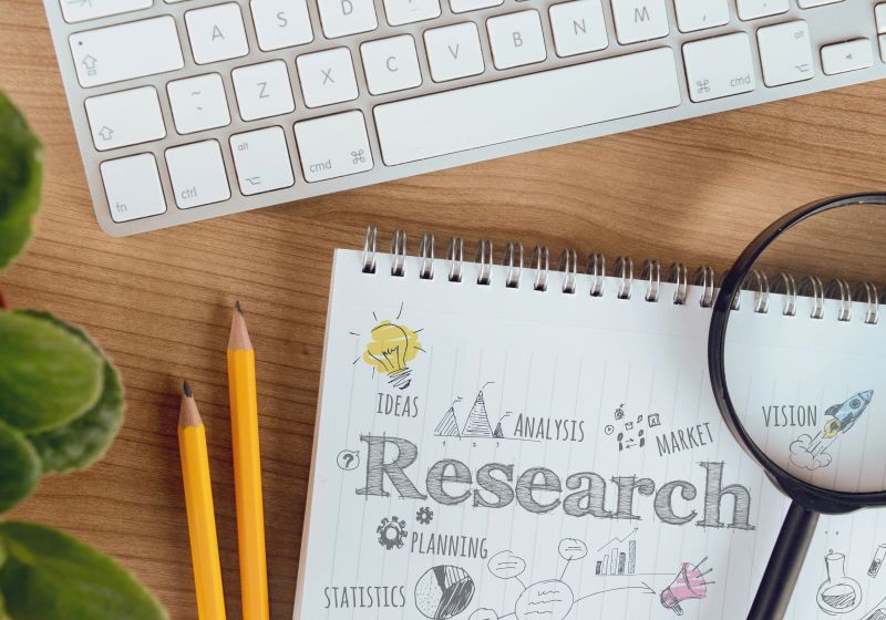The image shows a keyboard, paencils and magnifying glass with a paper pad depicting IBS Research