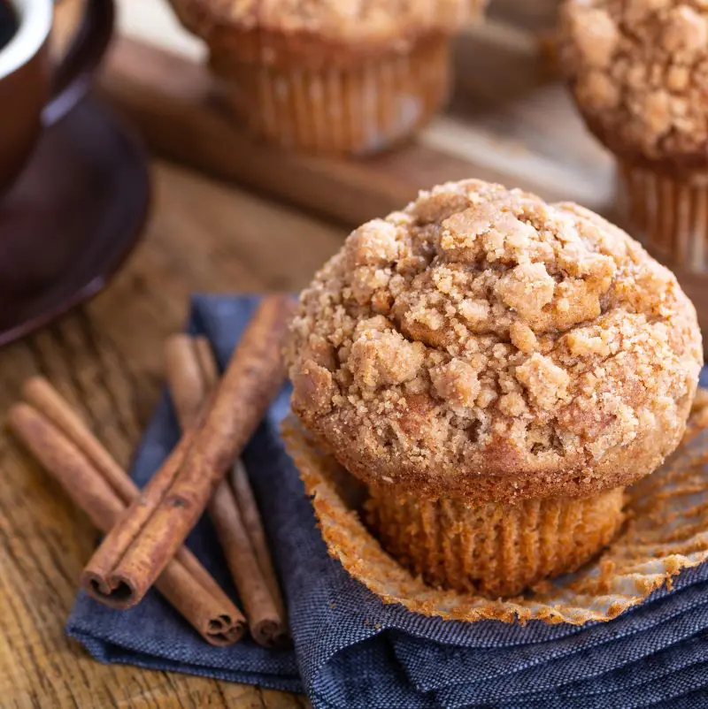 A Low FODMAP Cinnamon Muffin resting on a blue napkin with a cup of coffee and some cinnamon sticks nearby