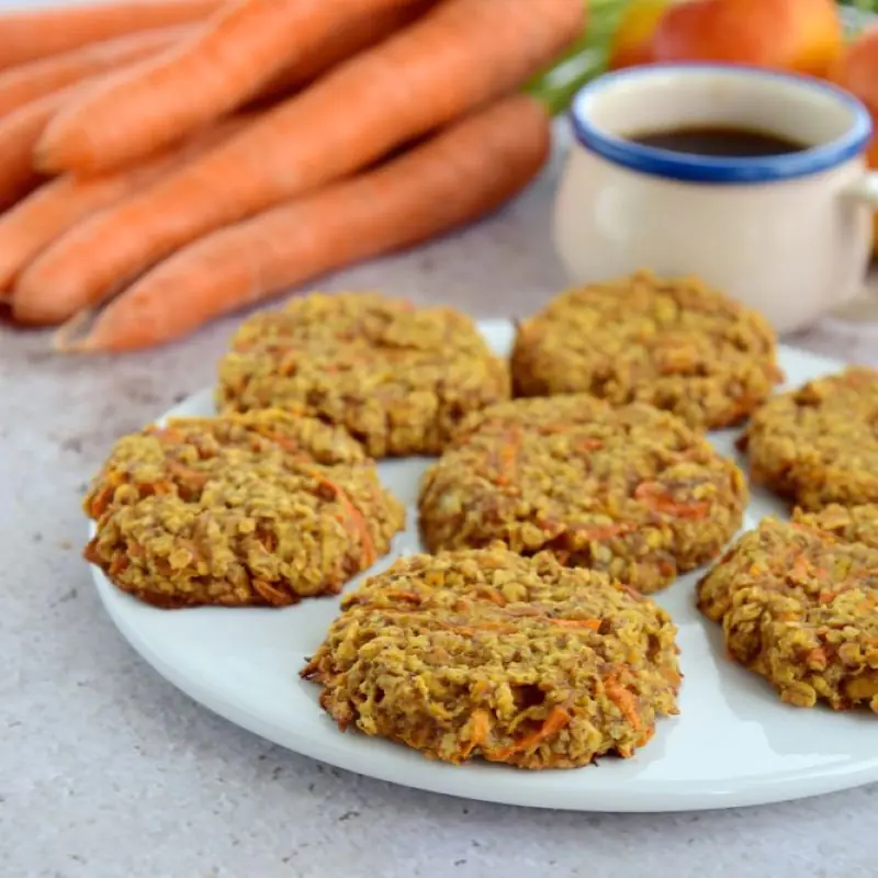 A plate showing some low FODMAP Carrot and Oak Cookies. In the background is a cup of coffee and a bunch of carrots